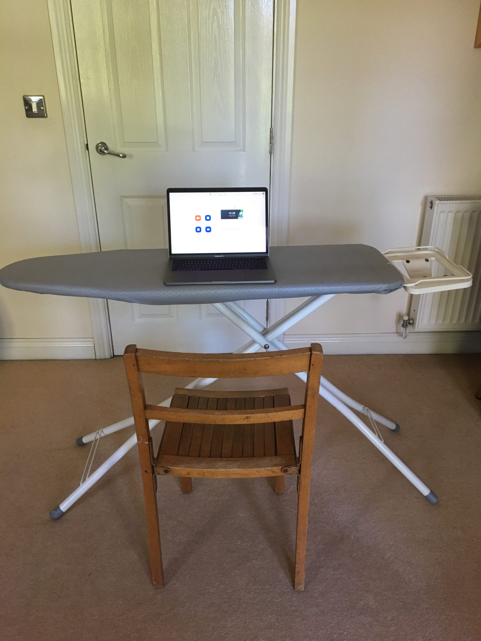 Working from home. Laptop open on an ironing board. Wooden chair in front of ironing board.